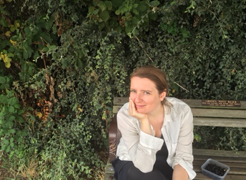 A girl is sitting on a bench in front of some bushes. She is smiling. 